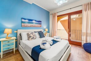 A hotel room with a bed, a nightstand with a lamp, a painting of a beach, and a balcony with curtains.