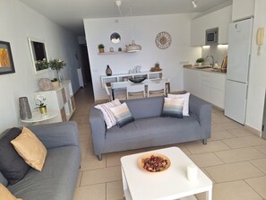 A modern living room with a grey sofa, a white coffee table, and a kitchen area with white cabinets and appliances.