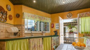 Une cuisine avec un mur recouvert de carreaux, une fenêtre ornée d’un rideau à motifs floraux et un saladier rempli d’oranges posé sur la table.