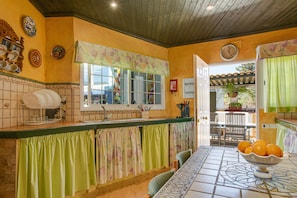 A kitchen with a tiled backsplash, a window with a floral curtain, and a bowl of oranges on the table.