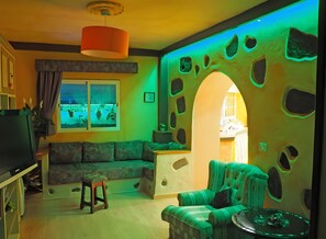 Een woonkamer met een groene bank, een groene fauteuil, een klein tafeltje, een televisie en een raam met uitzicht op planten.