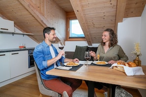 Zona pranzo con tavolo in legno, due persone sedute, piatti di spaghetti, bicchieri da vino e un vaso con fiori secchi.