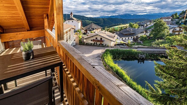 A wooden balcony with a view of a village, a pond, and mountains in the background. - Luxury Apartment "Weißhorn" with Mountain View and private Steam Sauna (Deutschnofen)