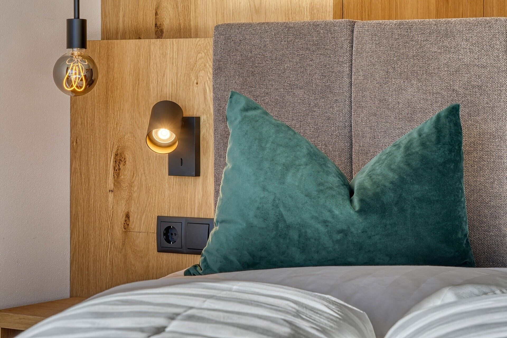 Un letto con una coperta bianca, un cuscino verde, un punto luce a parete e una presa elettrica.