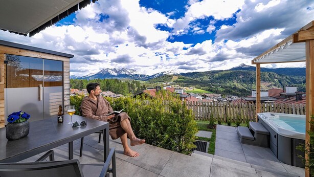 A man reading on a terrace with a mountain view, a hot tub, and a table with a beer bottle and glass. - Apartment "Schlossberg  Rosengarten" with Mountain View, Private Finnish Sauna & Garden (Deutschnofen)