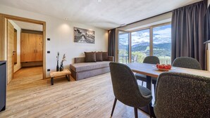 A modern room with a wooden floor, a sofa, a dining table with chairs, a painting on the wall, and a view of the outdoors. - Apartment "Schlossberg  Rosengarten" with Mountain View, Private Finnish Sauna & Garden (Deutschnofen)