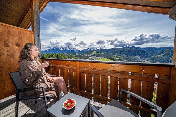 A person sitting on a balcony with a wooden railing, overlooking a mountainous landscape with a bowl of strawberries on a table.