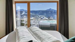 Ein Bett mit weißer Bettwäsche, Blick auf eine verschneite Berglandschaft und einen Poolbereich durch ein Fenster.