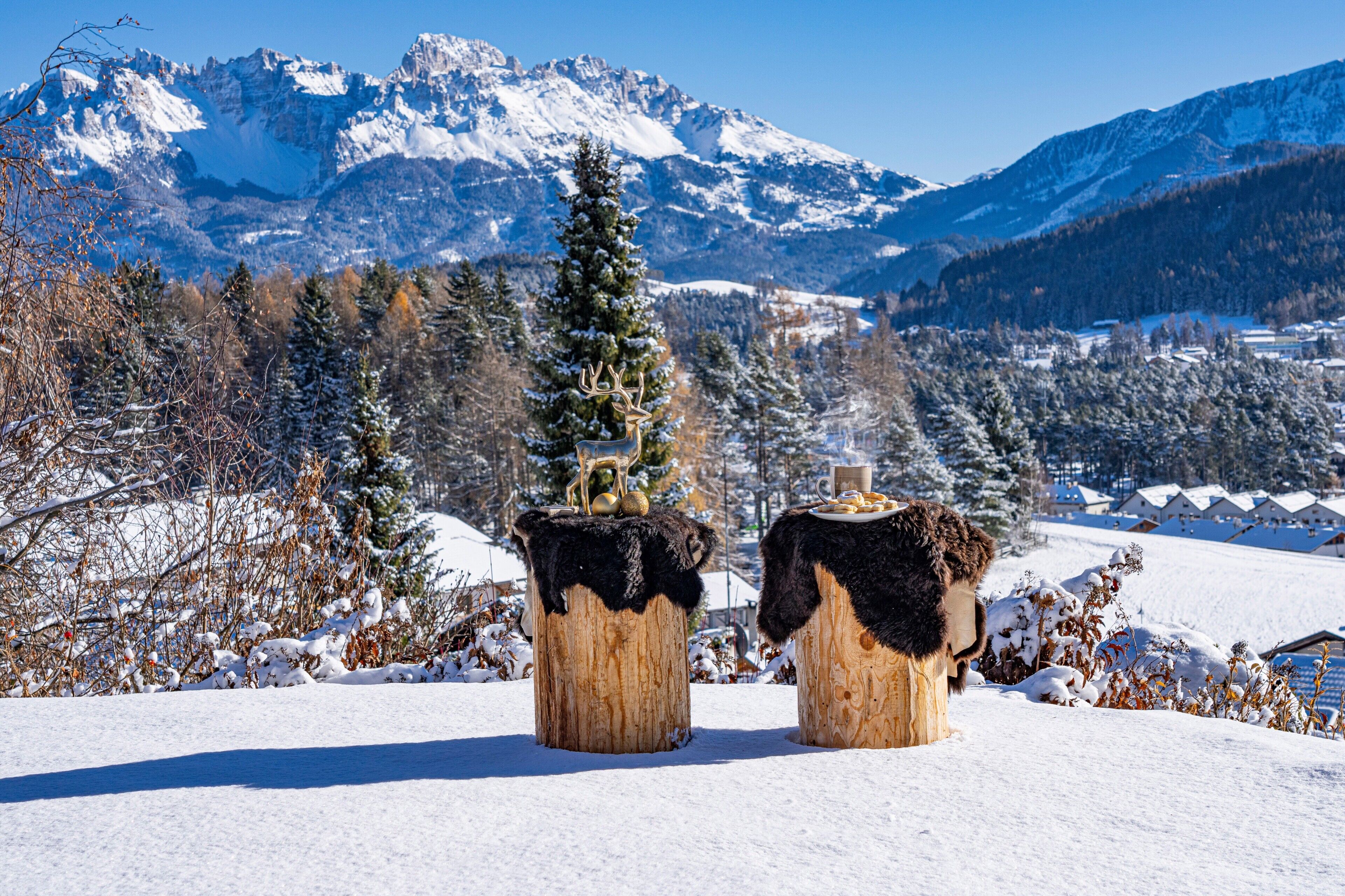 A snowy landscape with two wooden tables with fur throws, a pine tree, and a mountain range in the background.