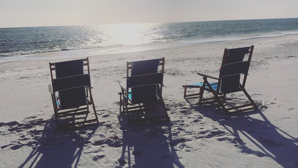 Drei Strandstühle stehen in einer Reihe auf sandigem Strand, Fußabdrücke führen zu ihnen.