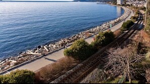 Vista costera con un paseo pavimentado al lado de una vía de tren, un puente a lo lejos y un litoral rocoso.