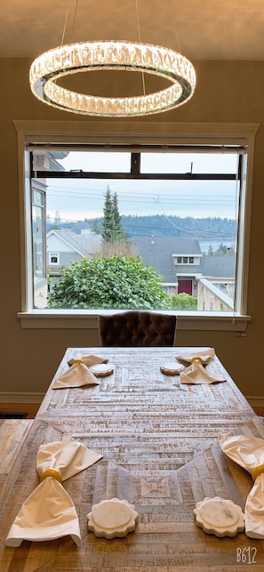 Enjoy a meal with a view of the surrounding neighborhood and forest from this dining area, featuring a chandelier, tufted leather chair, and a beautifully set table.