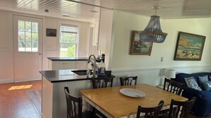 A kitchen with a wooden island, white cabinetry, and a dining area with a wooden table and chairs. There are two windows with blinds, a chandelier, and artwork on the walls. - Block Island The Way It Used To Be!!!  Steps From Everything!!! (New Shoreham)