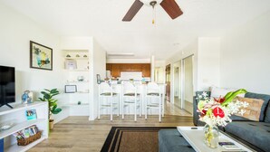 A modern living room with a flat-screen TV, a coffee table with a vase of flowers, a grey sofa, and a dining area with white chairs and a kitchen in the background. - Kona coast Ali'i Dr location by ocean, free, PKG WIFI salt water pool Poke shack (Kailua-Kona)