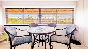 A balcony with a round table and four chairs, a fan, and a view of palm trees and buildings through the window. - Kona coast Ali'i Dr location by ocean, free, PKG WIFI salt water pool Poke shack (Kailua-Kona)