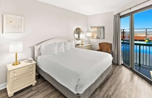 Habitación de hotel con una cama grande, una mesita de noche con lámpara, una silla, un balcón con vista al mar y un cuadro en la pared.