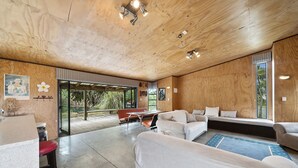 Un interior modern amb un sostre de fusta, zona d'estar amb una banca i sofàs, taula de dinar i una finestra gran amb vistes a vegetació.