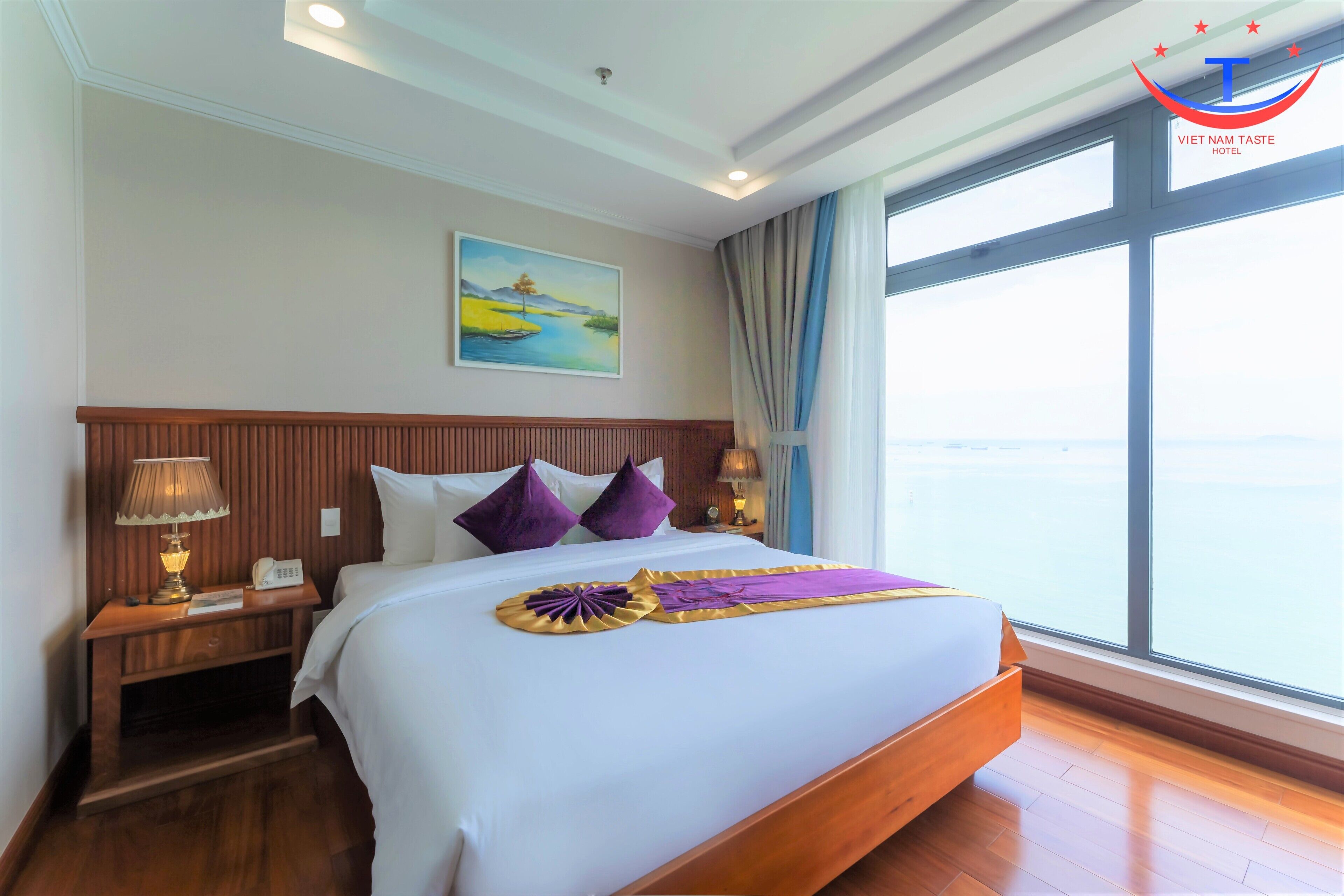 A hotel room with a large bed, a nightstand with a lamp, a painting on the wall, and a window with a view.