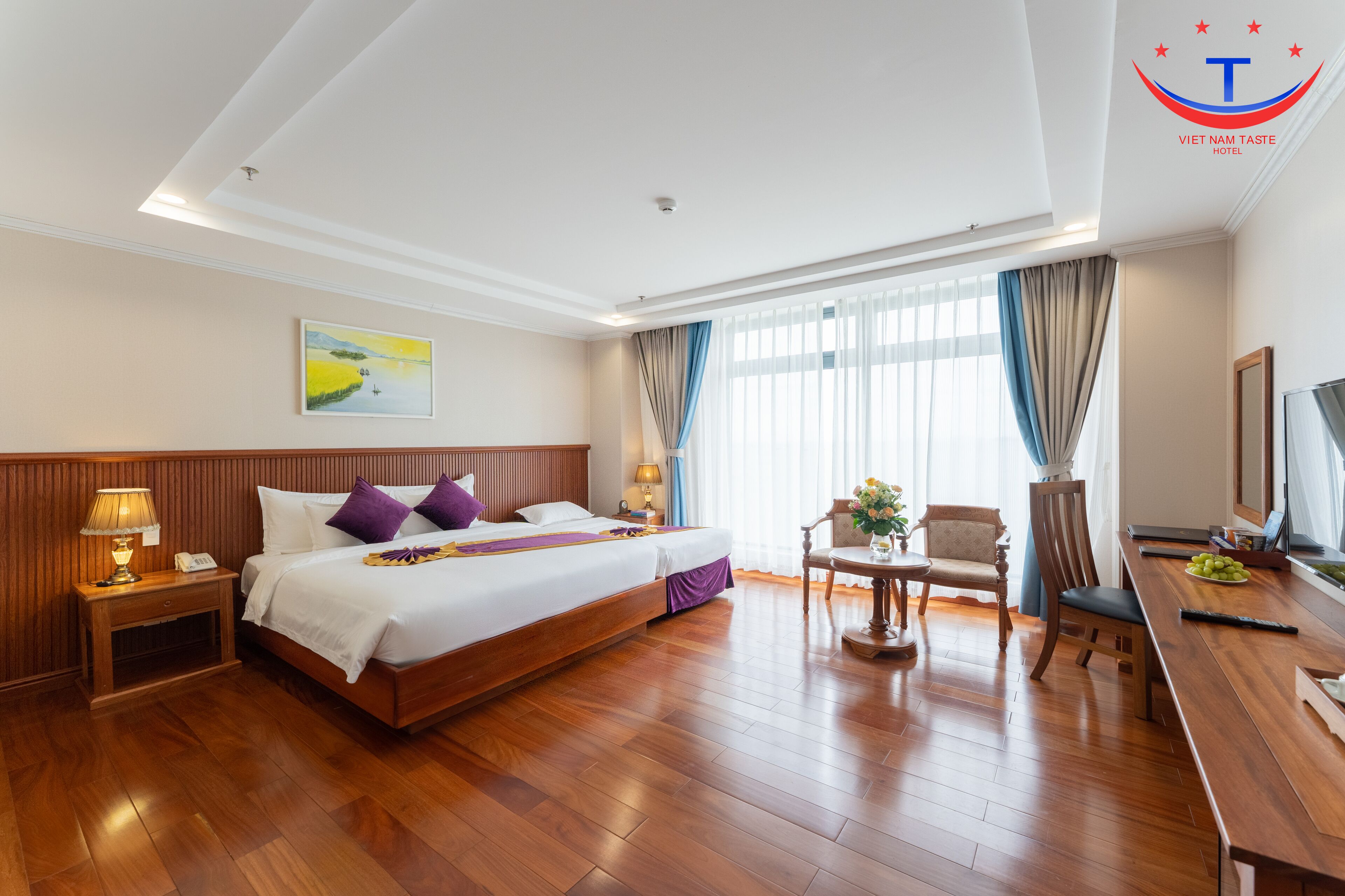 A hotel room with a large bed, wooden flooring, a desk with a chair, a television, and a painting on the wall.
