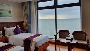 A hotel room with a large window offering a view of the sea, a bed with a purple and gold bedspread, a bedside table with a lamp, and a small round table with chairs. - Vietnam Taste Hotel Quy Nhon - Beachfront (Quy Nhon)