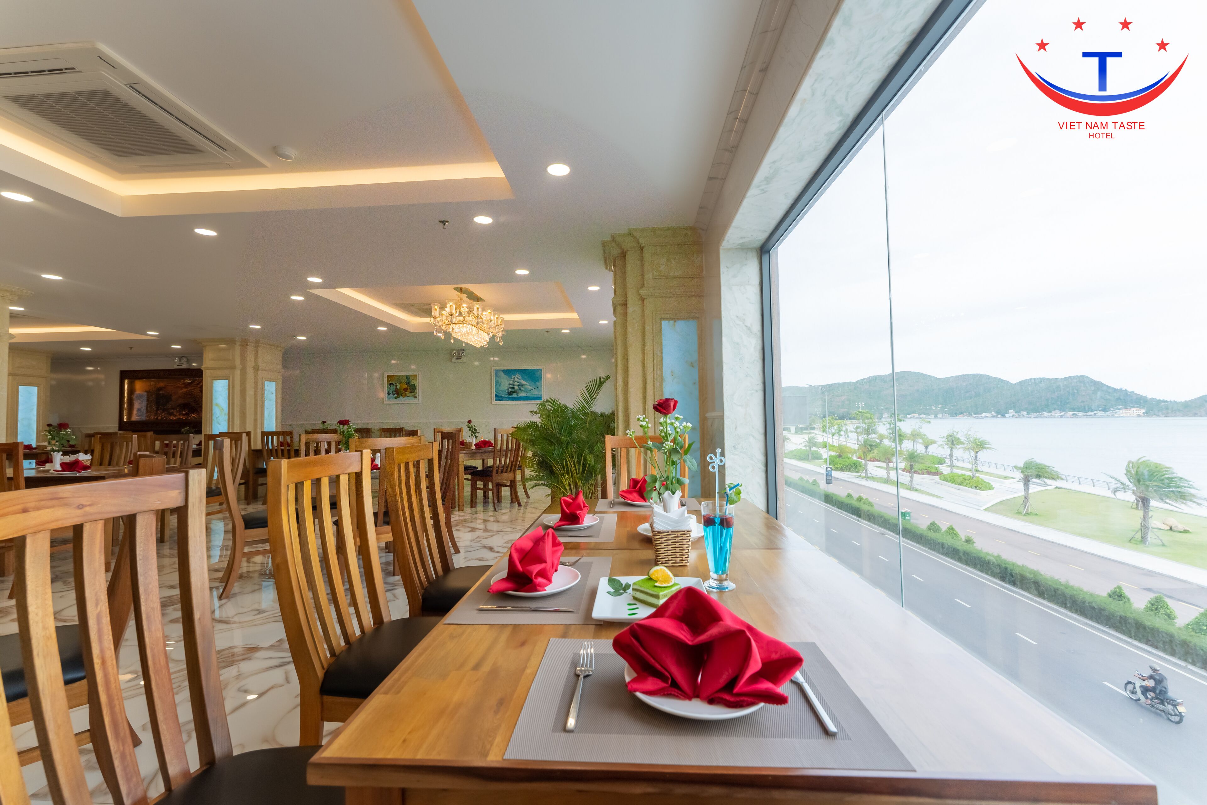 Un restaurant amb una taula llarga preparada per dinar, un lampadari i vista a un cos d'aigua.
