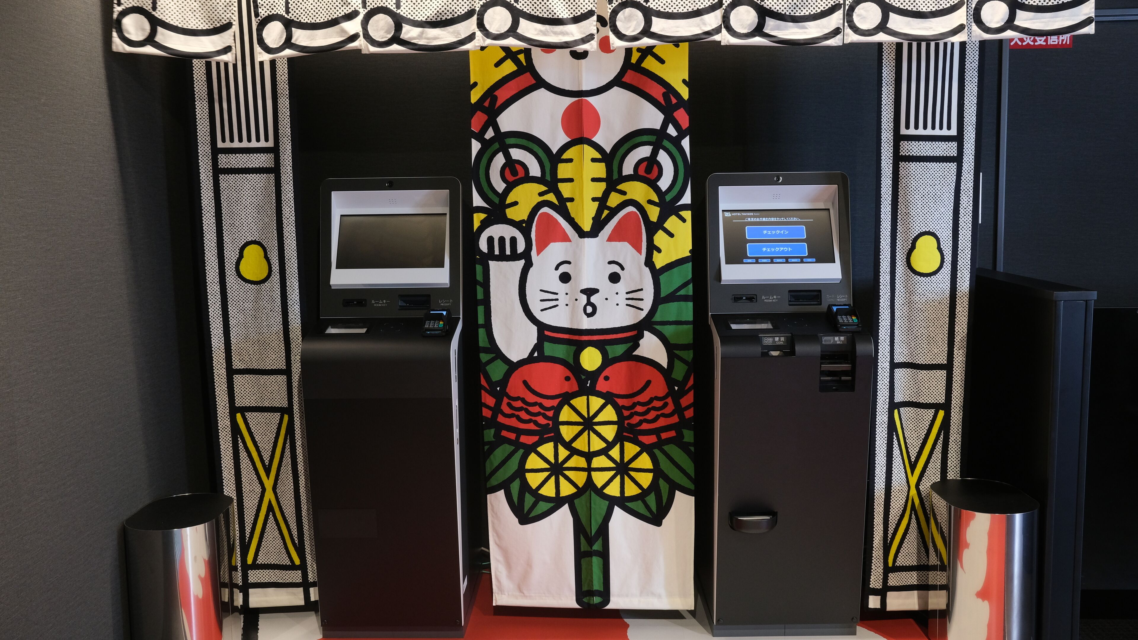 A hotel lobby with a large mural of a cat and a traditional Japanese design. There are two reception desks with digital displays and a small kiosk.
