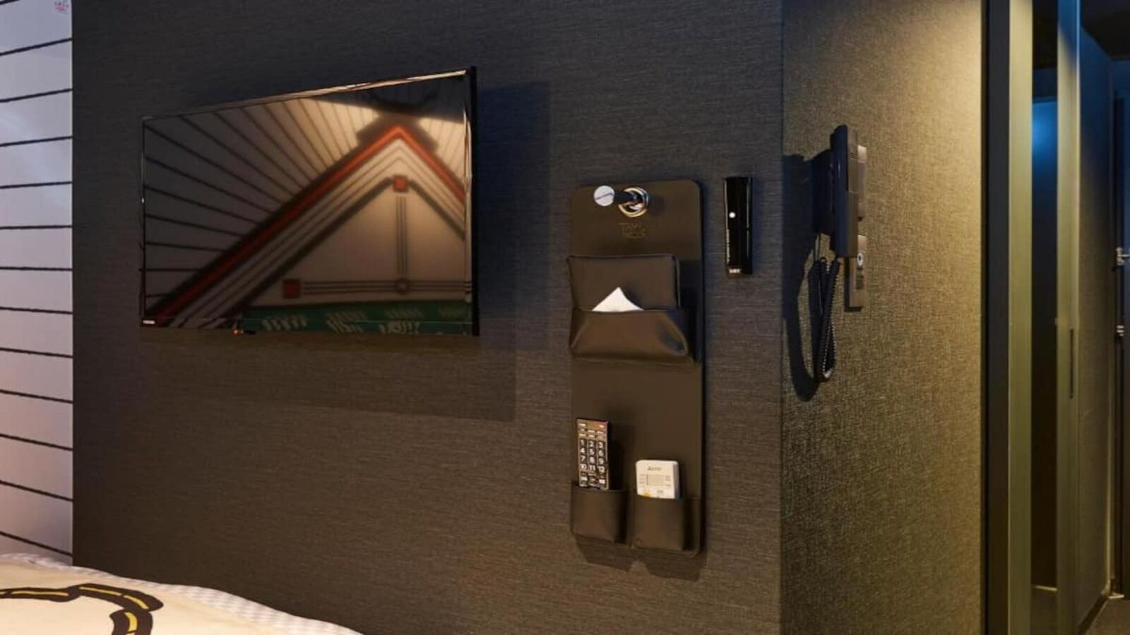 A hotel room with a bed, a wall-mounted control panel, a telephone, and a mirror reflecting a staircase.