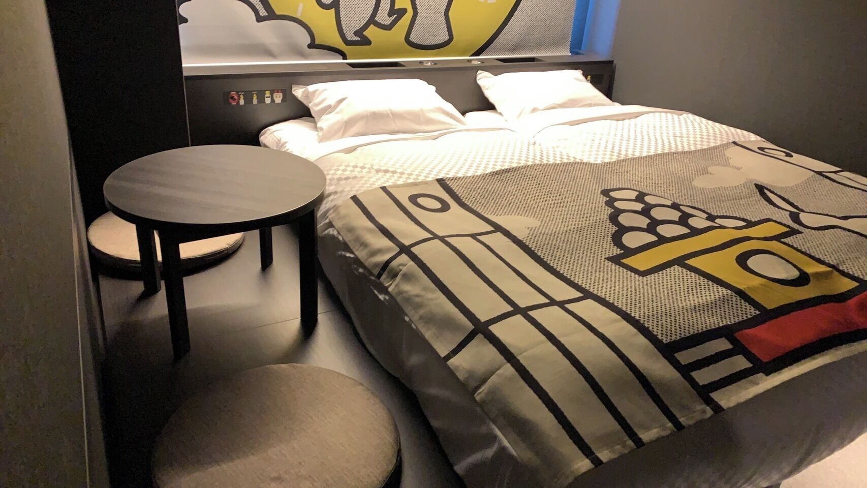 A compact hotel room with a bed, a round table, and a small stool.