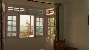 A bedroom with a bed, a wooden chair, a small table, and two windows with curtains. - Newly built bnb in a pristine village (Shimla)
