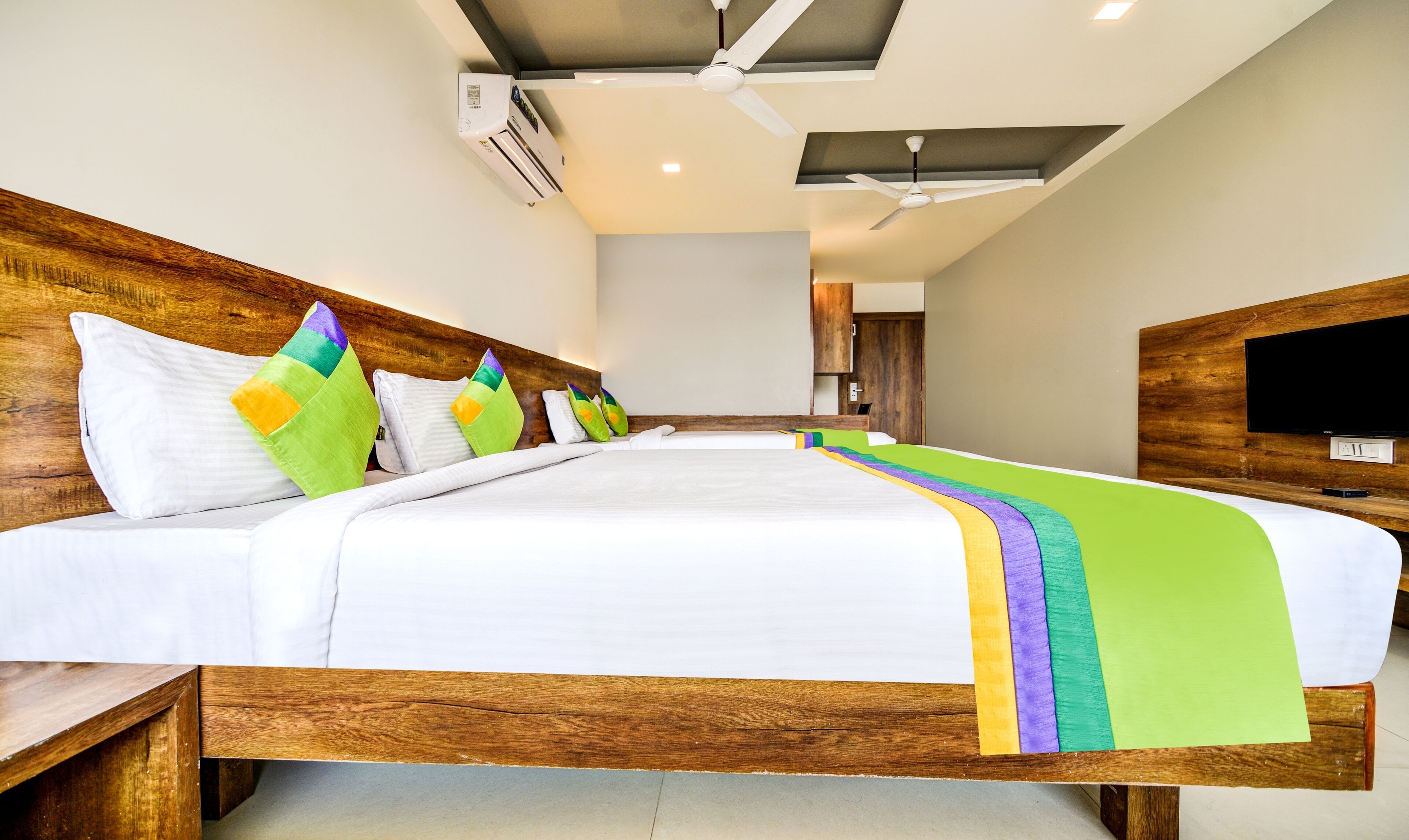 A hotel room with a large bed, wooden headboard, a flat-screen TV, and an air conditioning unit.