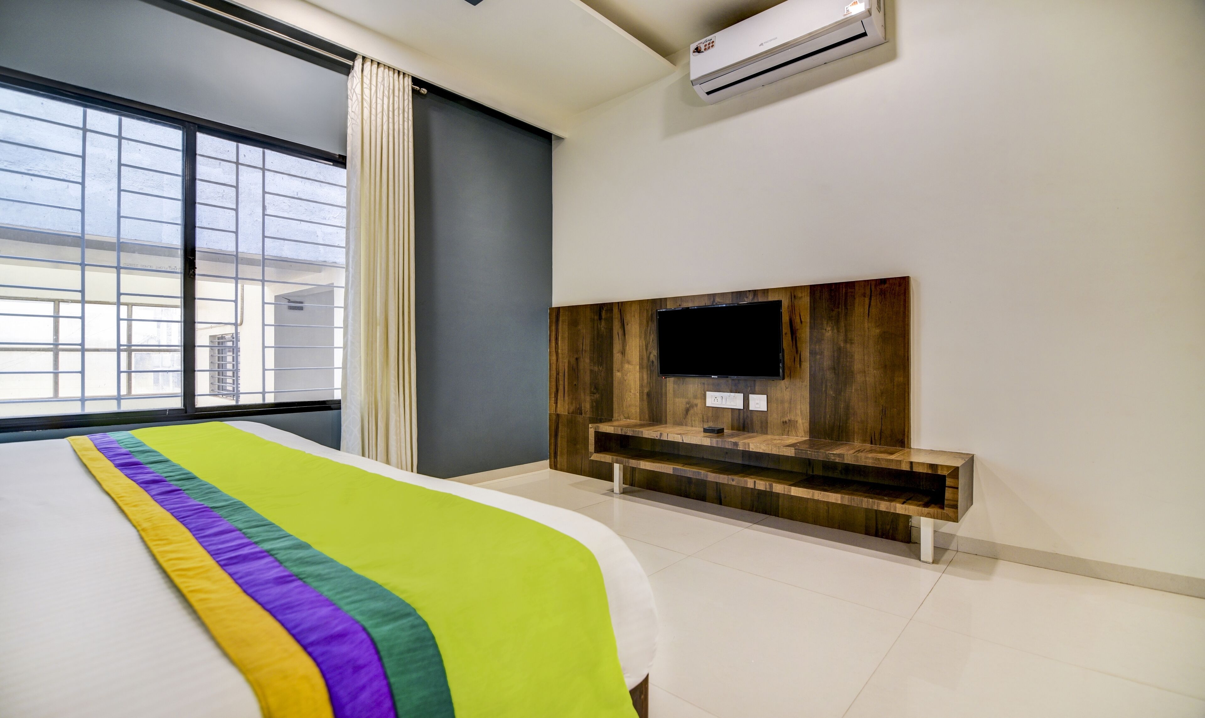A modern hotel room with a large bed, a mounted TV, and a window with blinds.