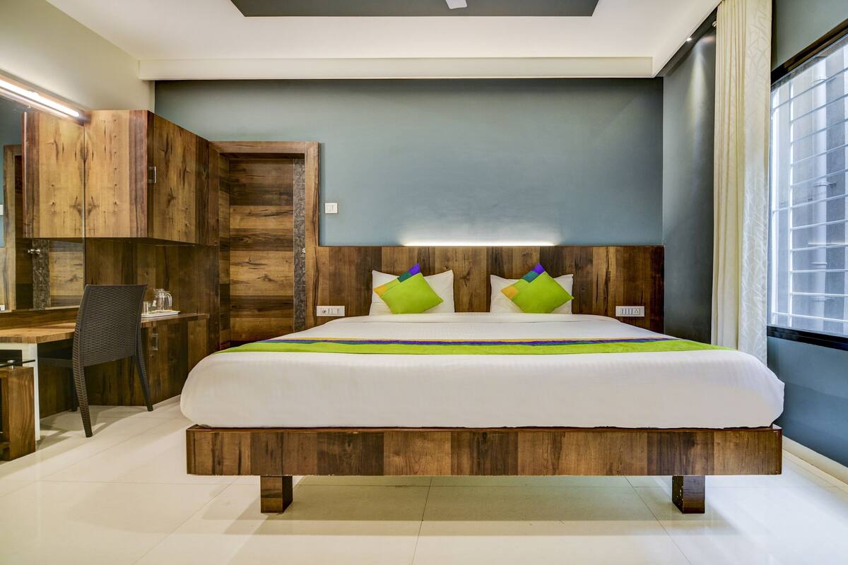 A modern bedroom with a wooden headboard, a large bed with green and purple pillows, a desk with a chair, and a window with blinds.
