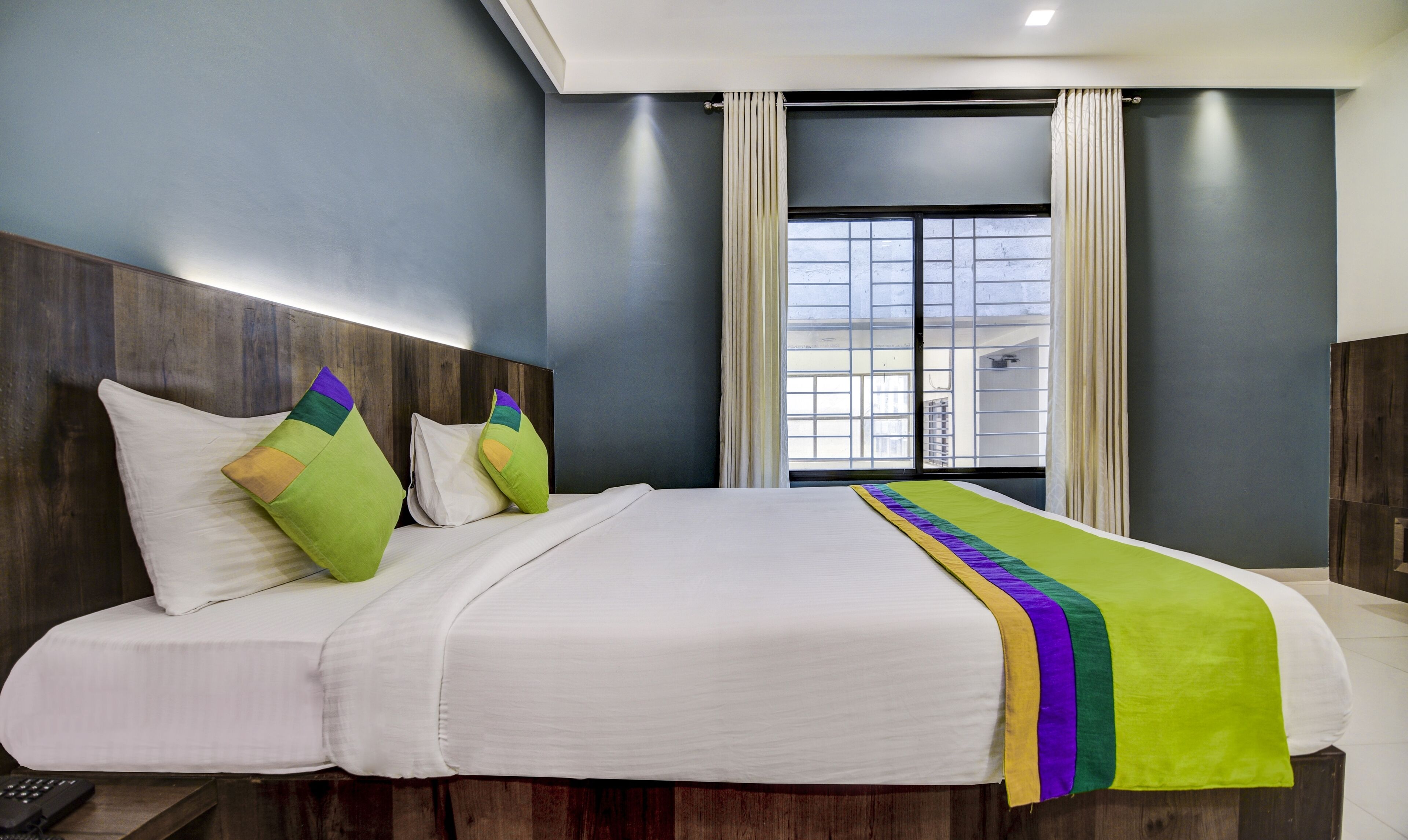 A hotel room with a large bed, a wooden headboard, a window with curtains, and a view of buildings outside.