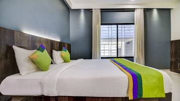 A hotel room with a large bed, a wooden headboard, a window with curtains, and a view of buildings outside.
