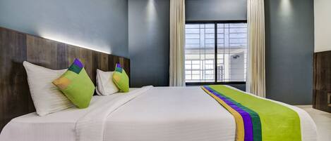 A hotel room with a large bed, a wooden headboard, a window with curtains, and a view of buildings outside.