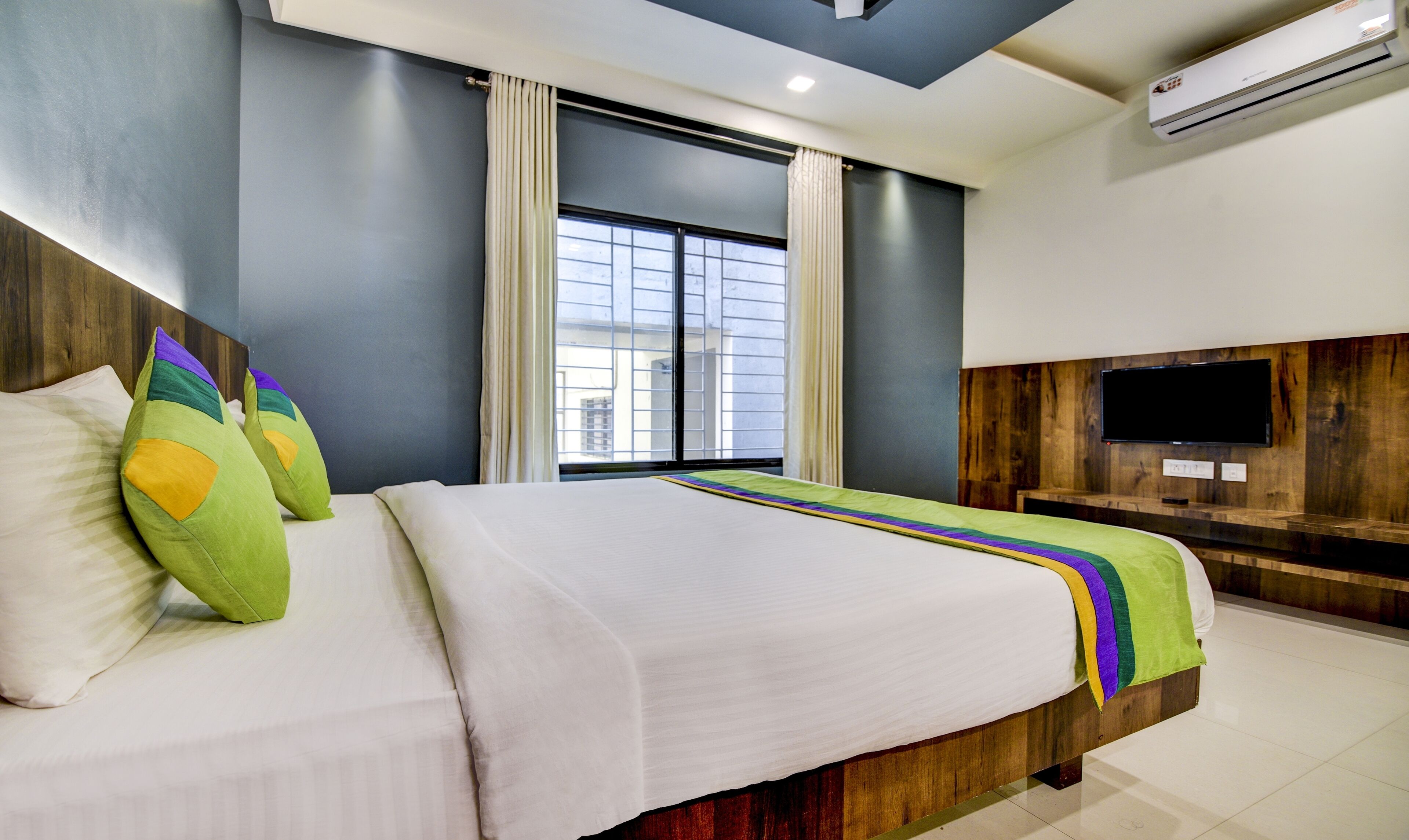 A modern hotel room with a large bed, a TV mounted on the wall, and a window with curtains.
