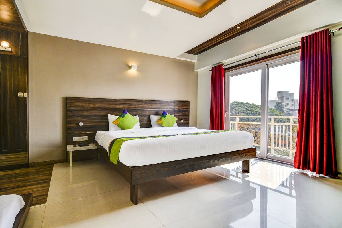 A hotel room with a large bed, a nightstand, a wooden headboard, a window with red curtains, and a balcony with a view.