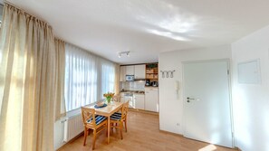 Quarto claro e bem iluminado com uma mesa de jantar e cadeiras, uma área de cozinha com armários e eletrodomésticos, e uma janela grande com cortinas.