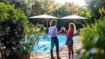 Zwei Personen stehen an einem Pool mit Sonnenschirmen und Grünpflanzen im Hintergrund.