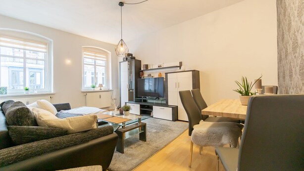 A modern living room with a sofa, coffee table, and TV stand. There is a dining area with a wooden table and chairs.