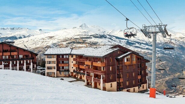 Ein Skigebiet mit mehrstöckigen Gebäuden, Skiliften und schneebedeckten Bergen im Hintergrund.