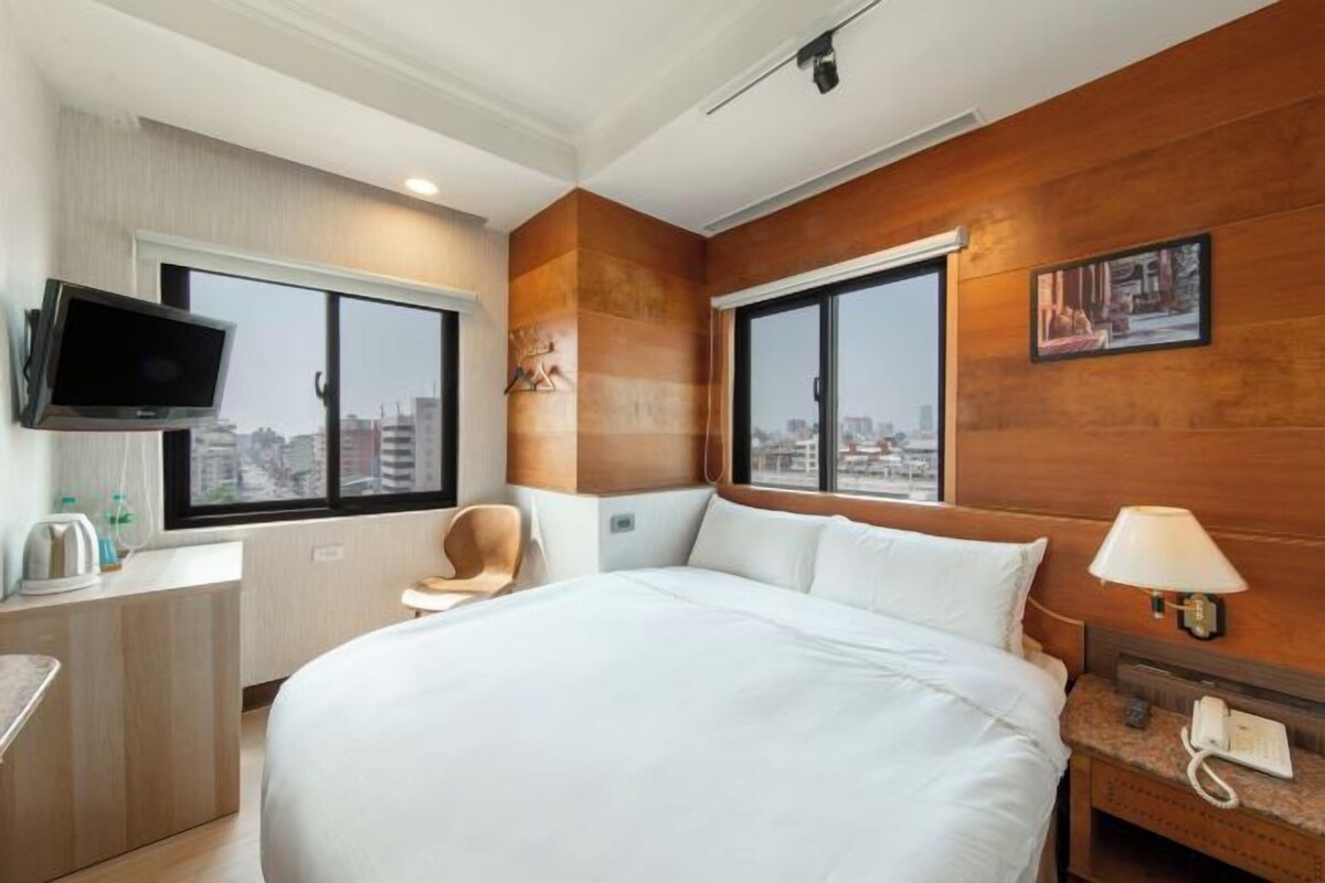 Ein Hotelzimmer mit einem großen Bett, einem an der Wand befestigten Fernseher, einem Schreibtisch mit Stuhl und einem Fenster mit Blick auf die Stadt.