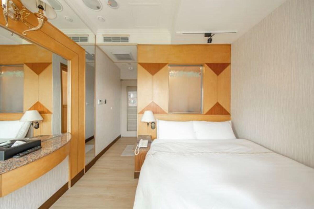 Ein Hotelzimmer mit einem großen Bett, einem Nachttisch mit Lampe, einem Spiegel und einem hölzernen Kopfteil.
