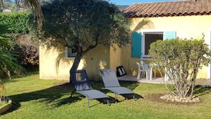 Un pequeño edificio amarillo con techo de tejas, un árbol en el patio y dos sillas de descanso.