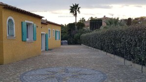 Un patio con pavimento de adoquines, un edificio amarillo con contraventanas turquesas y un seto.