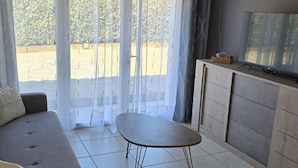 Una sala de estar con un sofá, una mesita, un televisor sobre un mueble y vista a un jardín a través de cortinas translúcidas.