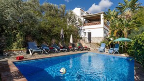 Una piscina con lettini e ombrelloni, circondata da vegetazione e un edificio con un balcone.