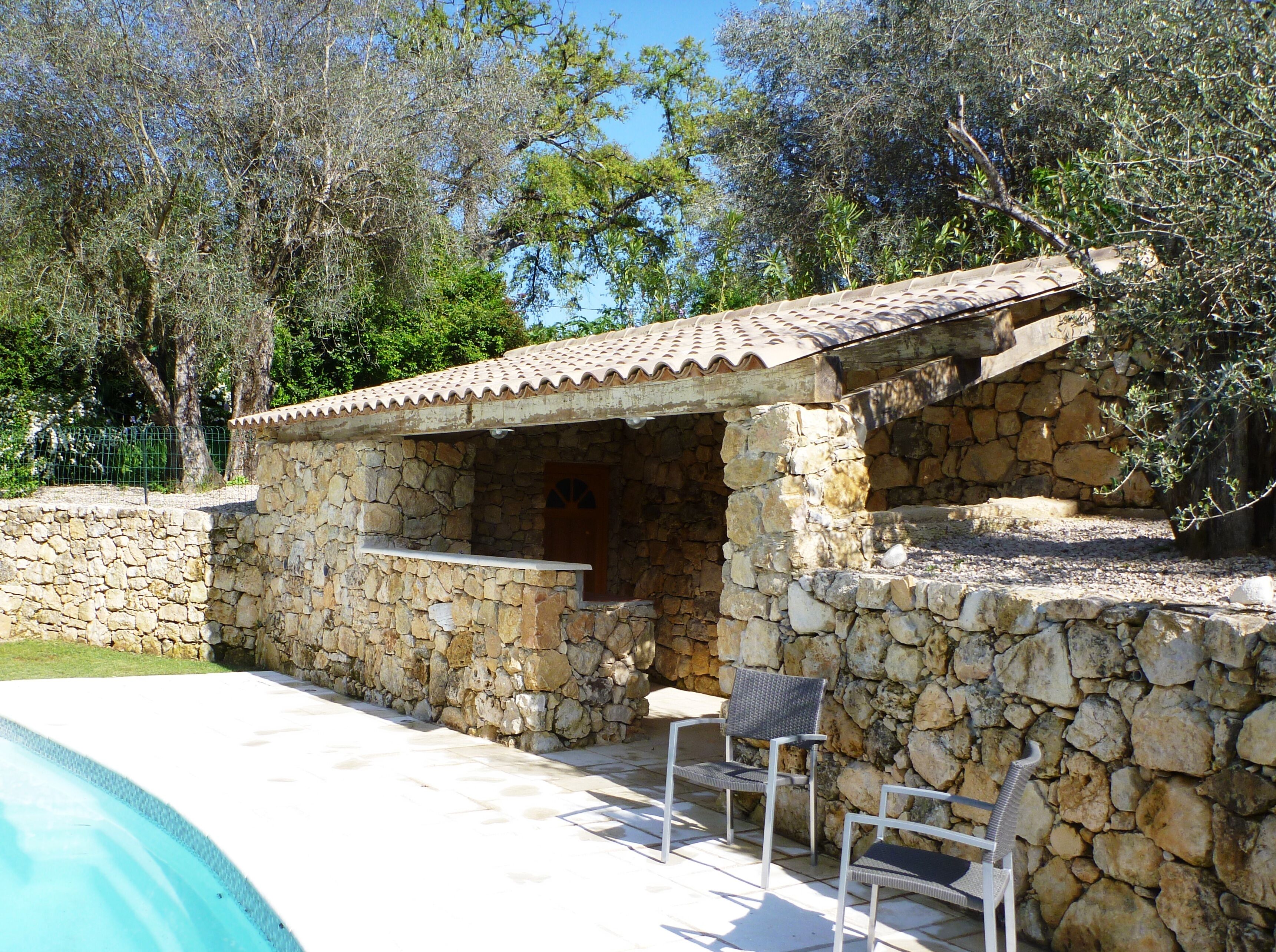 A stone building with a tiled roof, a pool, and two wicker chairs.