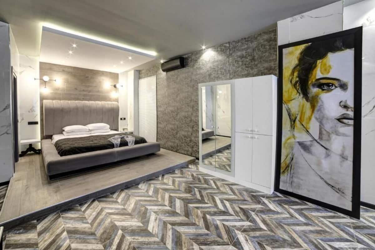 A modern bedroom with a large bed, a herringbone-patterned floor, a headboard with integrated lighting, and a large mirror cabinet.