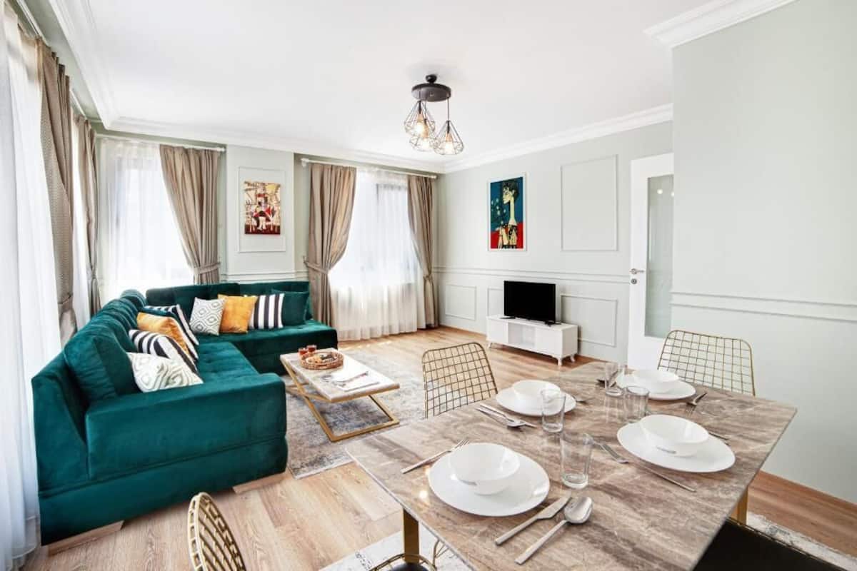 A modern living room with a green sofa, a dining table set for four, and a television in the corner.